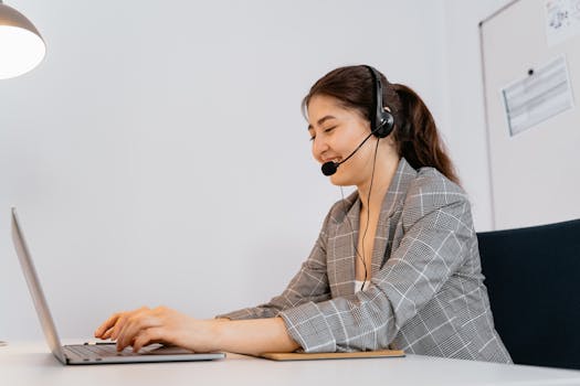 Woman in a plaid blazer working at a laptop with a headset in an office setting.